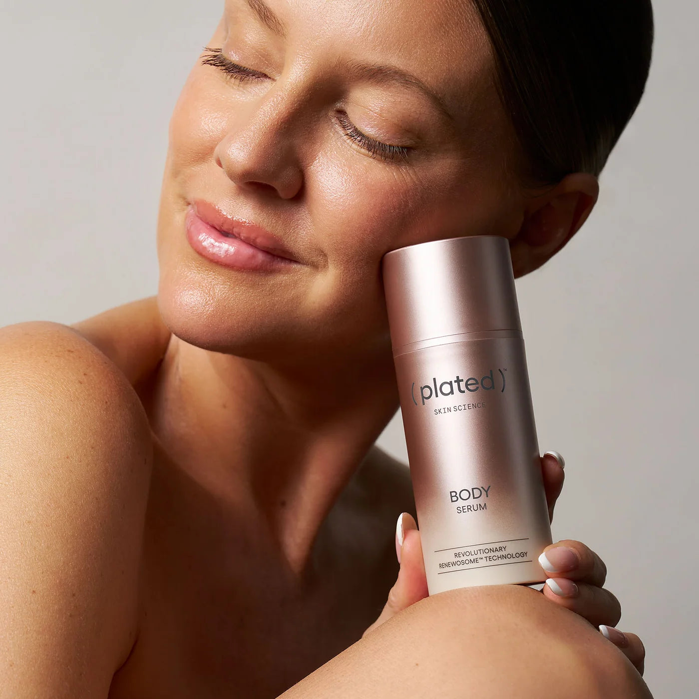 A woman holds a metallic rose gold bottle of (Plated) Skin Scents Body Serum against a neutral light gray background.
